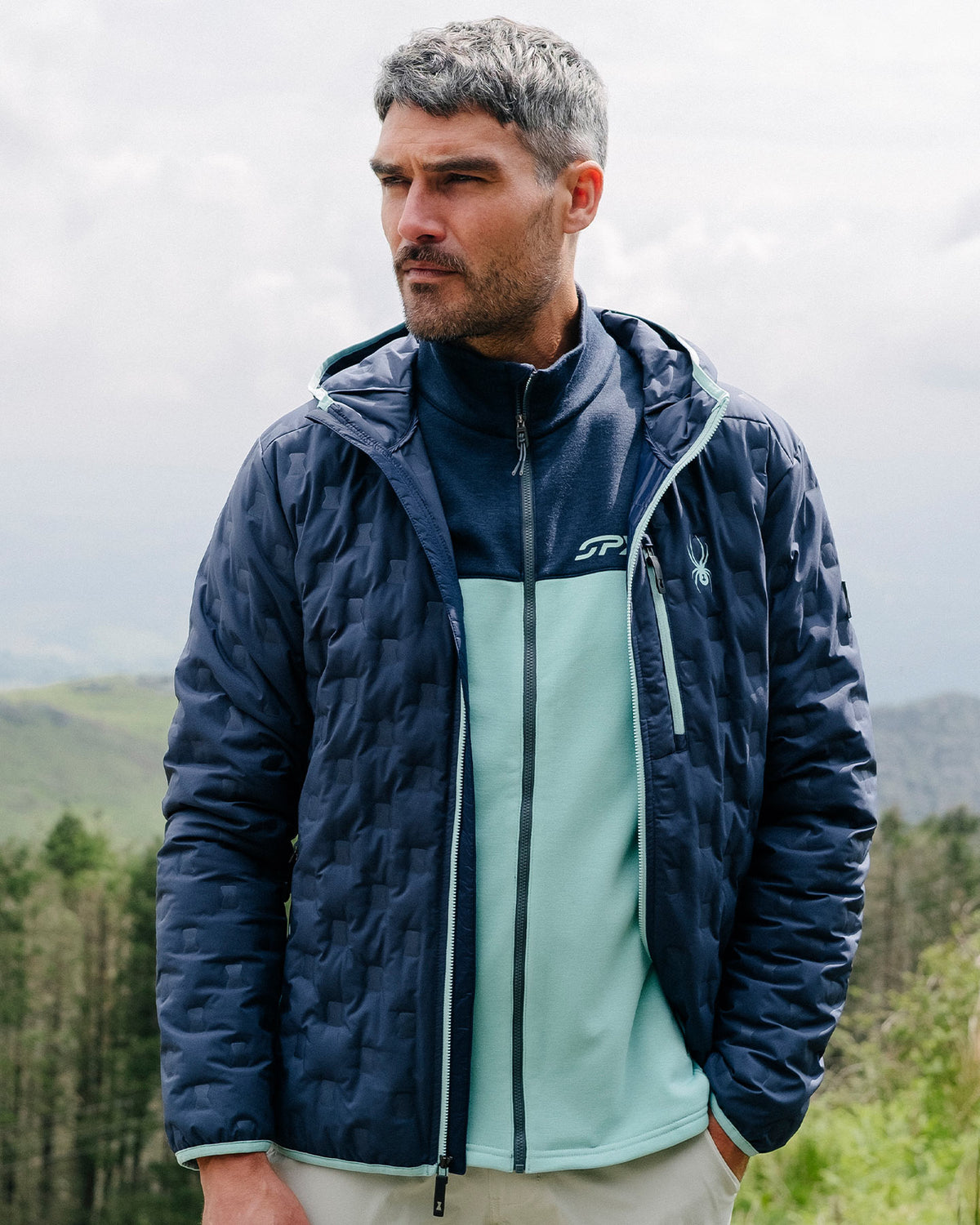 Man wearing a blue quilted jacket with a brand logo, standing outdoors with a forest in the background.