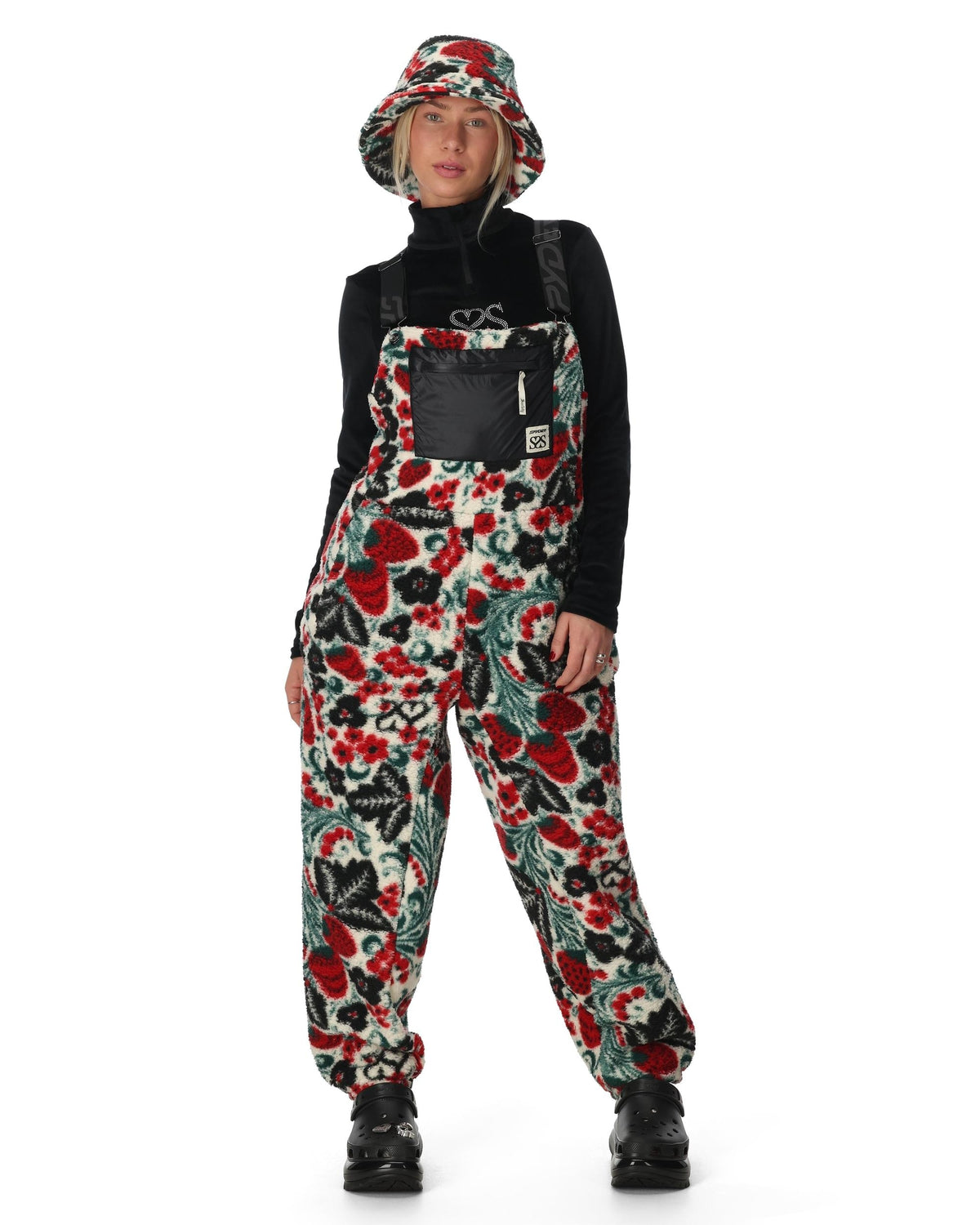 Person wearing a colorful floral patterned bib overalls with a black turtleneck and bucket hat on a white background.