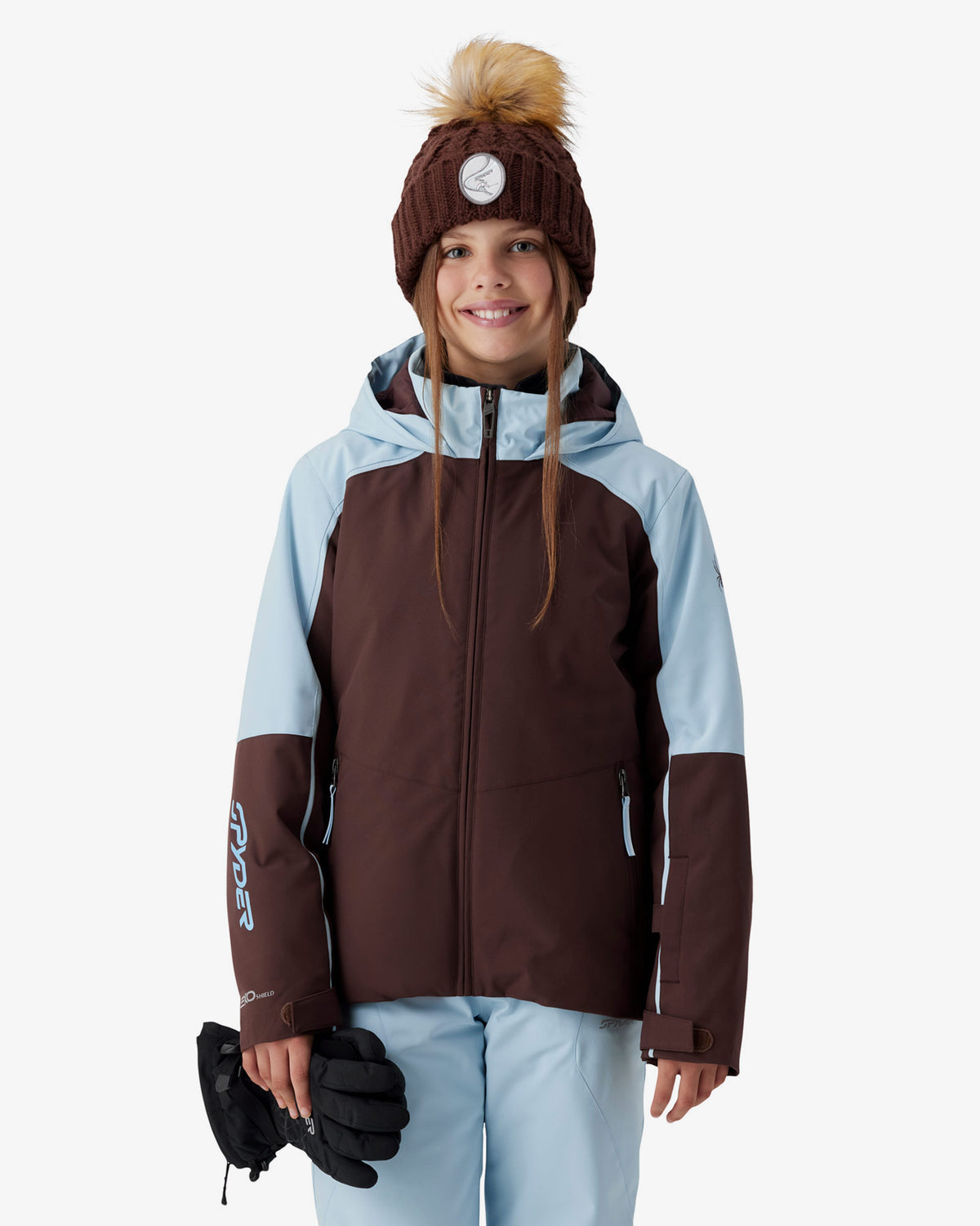 A young girl wearing a brown and light blue color-blocked ski jacket, a matching brown knit beanie with a tan pom-pom, and light blue ski pants, holding black gloves against a plain white studio background.