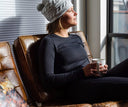Woman sitting on a brown leather couch holding a mug, wearing a Spyder base layer with gray knit hat.