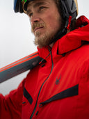 Person wearing a red Spyder jacket with a brand logo, holding skis against a snowy background