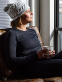 Woman sitting on a couch wearing a gray knit hat, wearing a base layer top and bottom, holding a white mug, looking out a window.