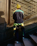 Person wearing a colorful outfit with a patterned hat and scarf, standing in front of an ornate wall.