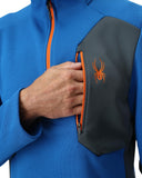 Blue jacket with orange zipper and black pouch featuring a logo, held by a hand.