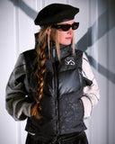 Person wearing a black puffer jacket with a brand logo, sunglasses, and a black beret against a white wall.