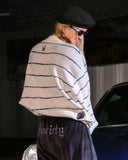 Person wrapped in a striped blanket with a logo, wearing sunglasses and a black cap, standing next to a car.