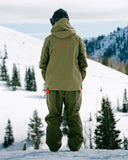 Person wearing a green snowsuit standing on a snowy landscape with trees and mountains.