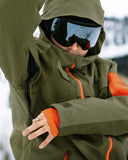 Person wearing a green jacket with orange accents and goggles, standing in a snowy landscape.