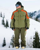 Person wearing a green and orange snowsuit standing in a snowy landscape with trees.