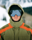 Person wearing a green and orange winter jacket with a hood and ski goggles in a snowy landscape.