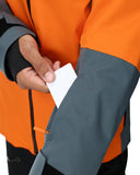 Person wearing an orange and gray jacket with a hand holding a white card.