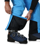 Person wearing blue pants and black ski boot with gaiters on a white background