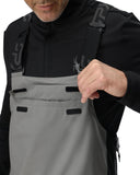 Person wearing a black jacket with a logo and gray bib overalls on a white background