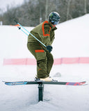 Skier in green and orange outfit performing a jump on a snowy course with trees in the background.