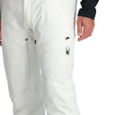 A man standing and wearing white ski pants with adjustable suspenders, a black shirt, and ski boots.