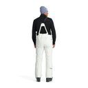 A man standing and wearing white ski pants with adjustable suspenders, a black shirt, and ski boots.