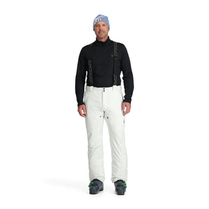 A man standing and wearing white ski pants with adjustable suspenders, a black shirt, and ski boots.
