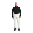 A man standing and wearing white ski pants with adjustable suspenders, a black shirt, and ski boots.